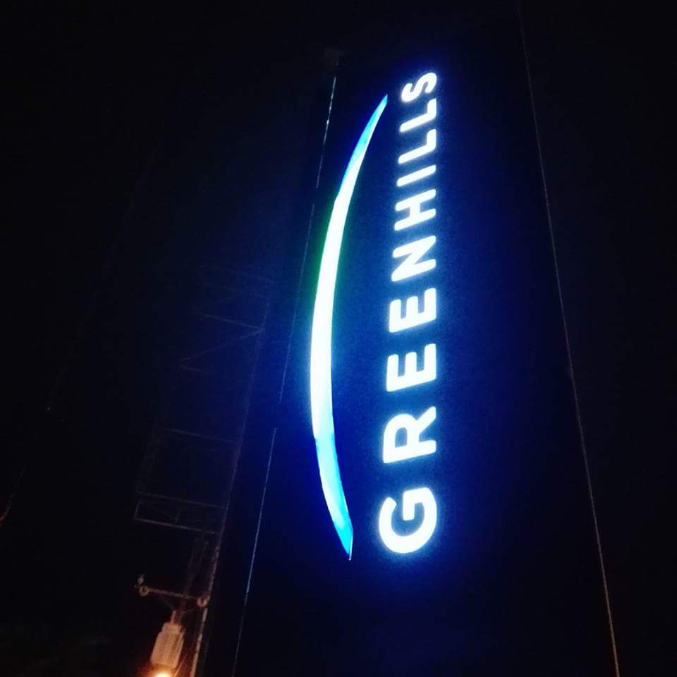 Vertical outdoor pylon signage for Greenhills in glowing white at night, seen in San Juan, by M&G Global Ads.